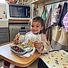 bags, bib, child, dishwasher, food, happy, high_chair, home_interior, indoor, kitchen, meal, microwave, pajamas, plate, smile, spoon, table, toddler, towels, window