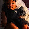 baby, child, infant, portrait, lying_down, black_outfit, hair_bow, bow, dress, tulle, earring, bed, pillow, bedding, indoor, close_up, face, serious_expression, warm_lighting, photography
