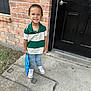 child, boy, smiling, standing, brick_wall, black_door, toy, blue_toy, casual_clothing, green_and_white_shirt, jeans, white_shoes, concrete_sidewalk, outdoor, happy, portrait, person, young_child, short_hair, daylight