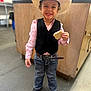 child, boy, cowboy_hat, vest, pink_shirt, jeans, belt, cookie, smile, standing, indoor, floor, wooden_cabinet, shoes, happy, portrait, casual, cute, young, person