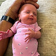 Susanna joined the competition — help win amazing prizes! baby, infant, pink_onesie, hand, watch, soft_surface, floral_pattern, skin, newborn, person, lying_down, cute, small, human, adult_hand, comfort, caretaking, resting, closeup, indoors