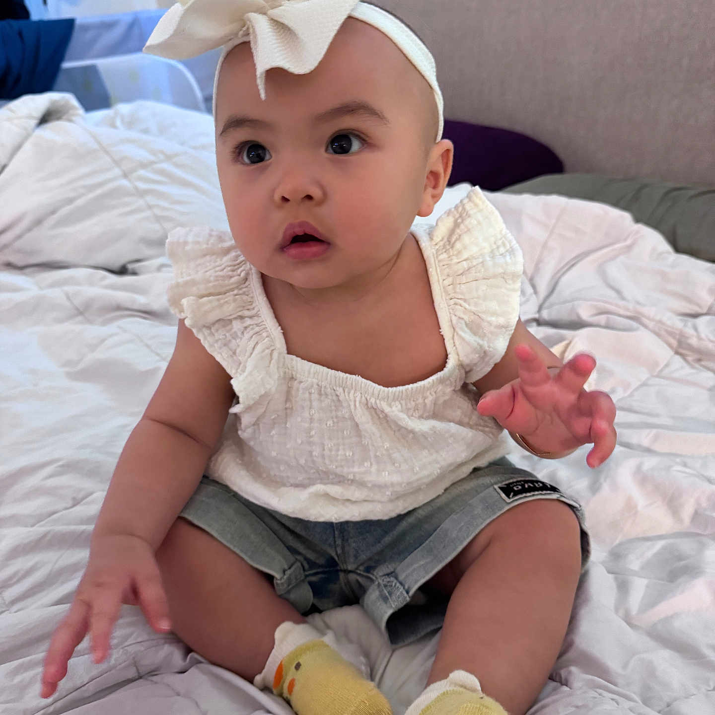 Harley is registered to the contest to win money with this photo: baby, child, infant, headband, bow, white_top, denim_shorts, yellow_socks, duck_design, bed, blanket, indoor, cute, curious, sitting, hand, face, eyes, person, clothing