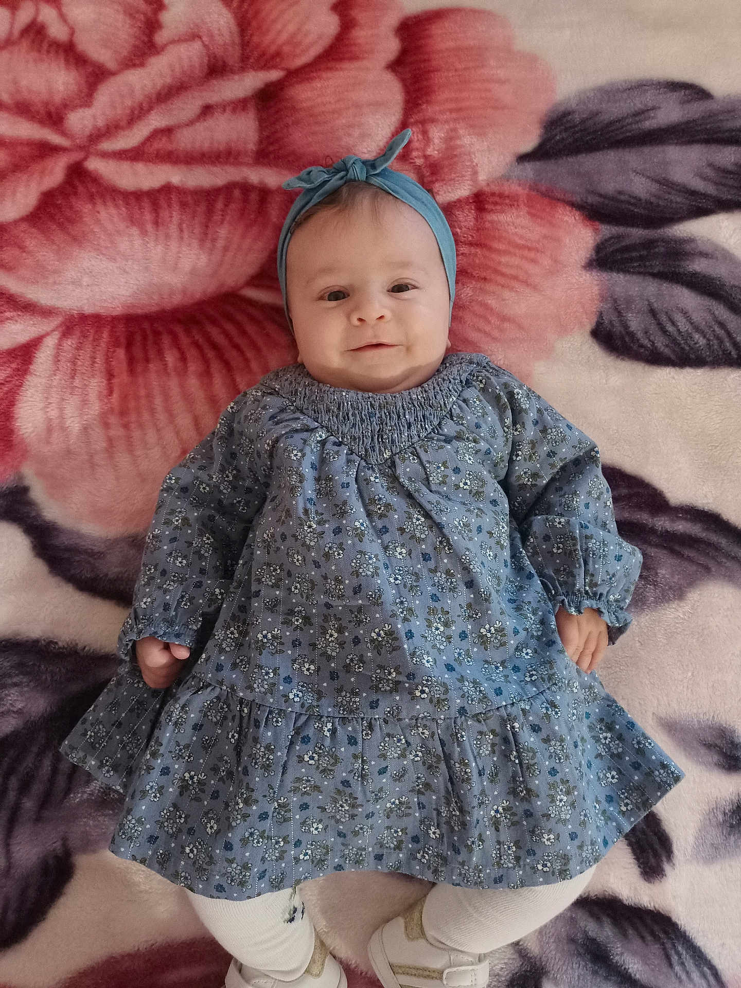 Sinda participe au concours pour gagner de l'argent avec cette photo : baby, infant, blue_dress, headband, blanket, floral_pattern, smiling, lying_down, white_tights, white_shoes, cute, child, portrait, indoors, soft_texture, happy, small_hands, cozy, comfortable, baby_fashion