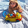 baby, child, hoodie, toy, toy_bus, animal_figures, blanket, outdoor, grass, happy, smiling, cute, playtime, sunny, infant, clothing, person, blue_blanket, yellow_toy, colorful