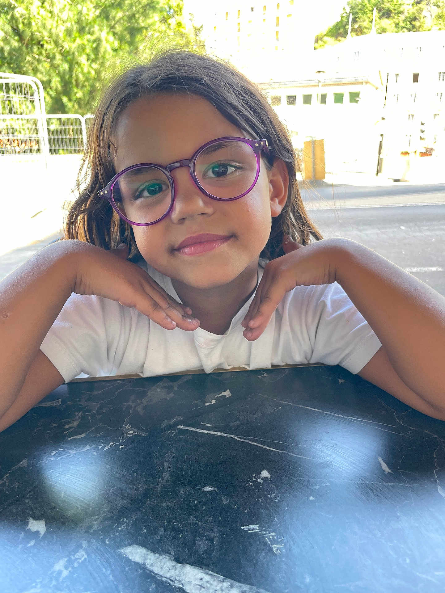 Yasmine a rejoint le concours — aidez-le/la à gagner de superbes lots ! child, glasses, purple_glasses, smiling, portrait, outdoor, table, marble_table, short_sleeve_shirt, white_shirt, happy, face, hands, posing, nature, daylight, person, casual, young, cute