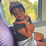 Yasmine a rejoint le concours — aidez-le/la à gagner de superbes lots ! child, girl, tiara, train, window, seat, purple_seat, shorts, black_tshirt, smile, pose, hand_gesture, nature_outside, trees, sunlight, casual_clothing, portrait, indoor, travel, young