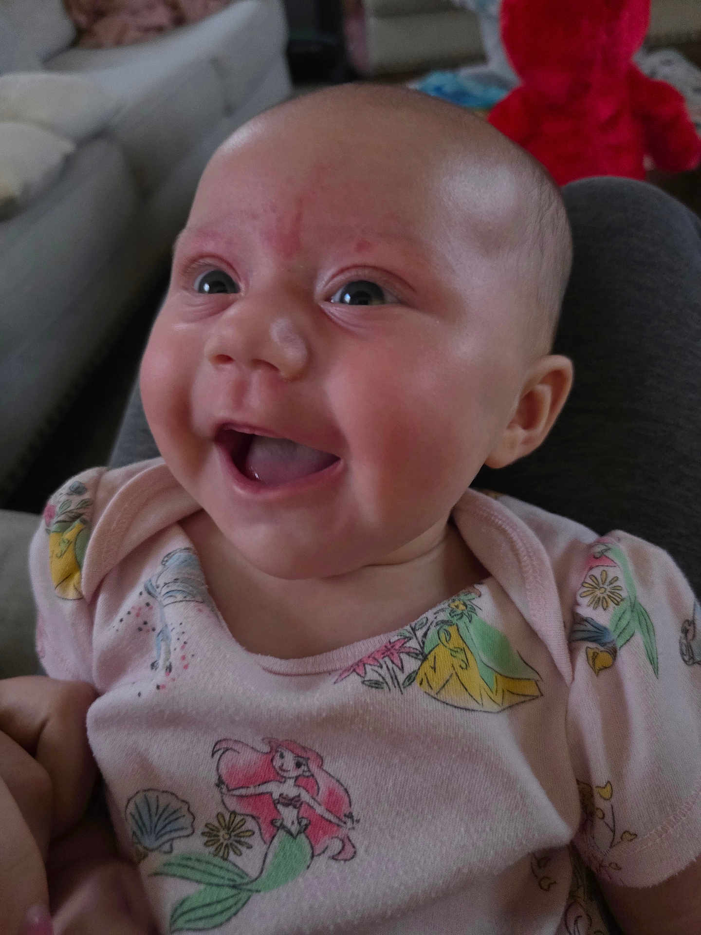 Mia is registered to the contest to win money with this photo: baby, smiling, indoor, child, face, clothing, pink_shirt, mermaid_pattern, happy, cute, person, infant, portrait, sitting, plush_toy, background, couch, cozy, head, eyes