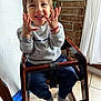 Kyrie is registered to the contest to win money with this photo: toddler, child, smiling, high_chair, wooden_chair, indoor, happy, sweatshirt, pants, socks, bracelet, hands, face, portrait, floor_tiles, curtain, brick_wall, natural_light, playful, cozy