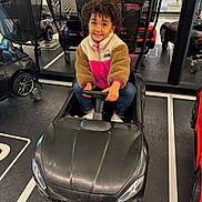 Ilyana a rejoint le concours — aidez-le/la à gagner de superbes lots ! child, toy_car, indoor, curly_hair, fleece_jacket, black_car, small_car, shopping_cart, floor_markings, happy, person, jeans, white_shoes, steering_wheel, parking_lot, smiling, play, vehicle, kid, casual_clothing