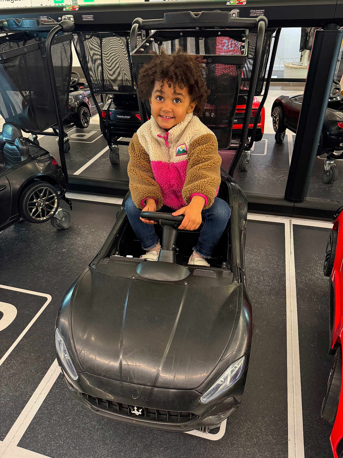 Ilyana a rejoint le concours — aidez-le/la à gagner de superbes lots ! child, toy_car, indoor, curly_hair, fleece_jacket, black_car, small_car, shopping_cart, floor_markings, happy, person, jeans, white_shoes, steering_wheel, parking_lot, smiling, play, vehicle, kid, casual_clothing
