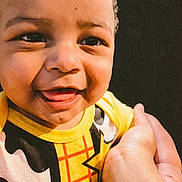 Major is registered to the contest to win money with this photo: baby, child, infant, smiling, happy, portrait, close_up, face, eyes, teeth, curly_hair, yellow_clothing, costume, superhero, being_held, adult_hand, arm, indoor, dark_background, photography