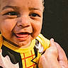 baby, child, infant, smiling, happy, portrait, close_up, face, eyes, teeth, curly_hair, yellow_clothing, costume, superhero, being_held, adult_hand, arm, indoor, dark_background, photography