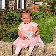 Lina a rejoint le concours — aidez-le/la à gagner de superbes lots ! toddler, child, pink_dress, doll, outdoor, grass, stone_wall, brick_house, greenery, sunlight, smiling, cute, toy, baby, person, garden, shoes, hair_clip, nature, portrait