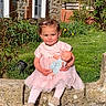 toddler, child, pink_dress, doll, outdoor, grass, stone_wall, brick_house, greenery, sunlight, smiling, cute, toy, baby, person, garden, shoes, hair_clip, nature, portrait