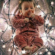 Louna participe au concours pour gagner de l'argent avec cette photo : baby, infant, onesie, pink_clothing, fairy_lights, blanket, cozy, cute, child, indoors, portrait, smiling, feet, hands, face, soft_lighting, warm_tones, sleepwear, resting, toddler
