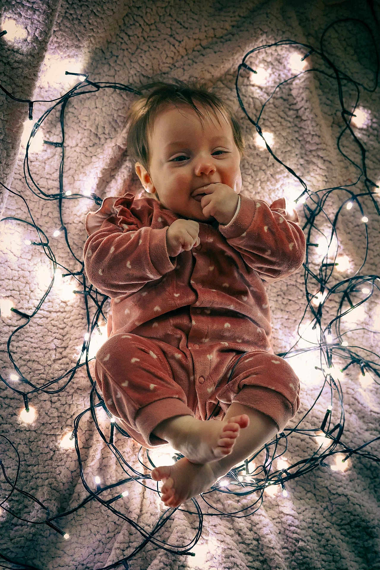 Louna participe au concours pour gagner de l'argent avec cette photo : baby, infant, onesie, pink_clothing, fairy_lights, blanket, cozy, cute, child, indoors, portrait, smiling, feet, hands, face, soft_lighting, warm_tones, sleepwear, resting, toddler