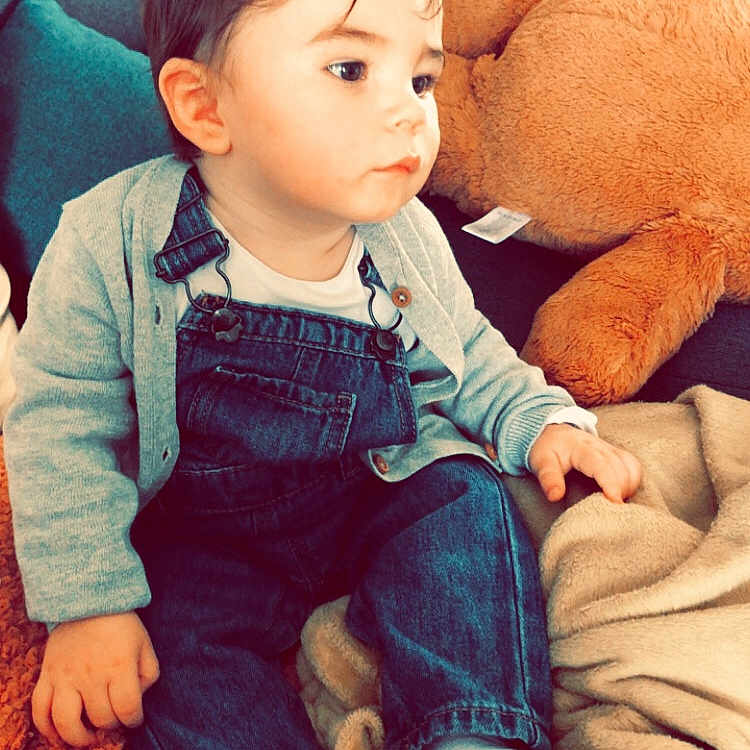 Wyley participe au concours pour gagner de l'argent avec cette photo : blanket, cardigan, casual_clothing, child, curly_hair, cute, denim_overalls, floor_tiles, home, indoor, pillow, plush_toy, portrait, relaxed, seated, socks, sofa, soft_toys, toddler, wooden_stool