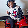 Jamarcus is registered to the contest to win money with this photo: child, boy, hoodie, spiderman, toy_vehicle, red, indoor, smiling, playful, hand_gesture, black_pants, floor, wall, furniture, small_vehicle, casual_clothing, young, portrait, happy, fun