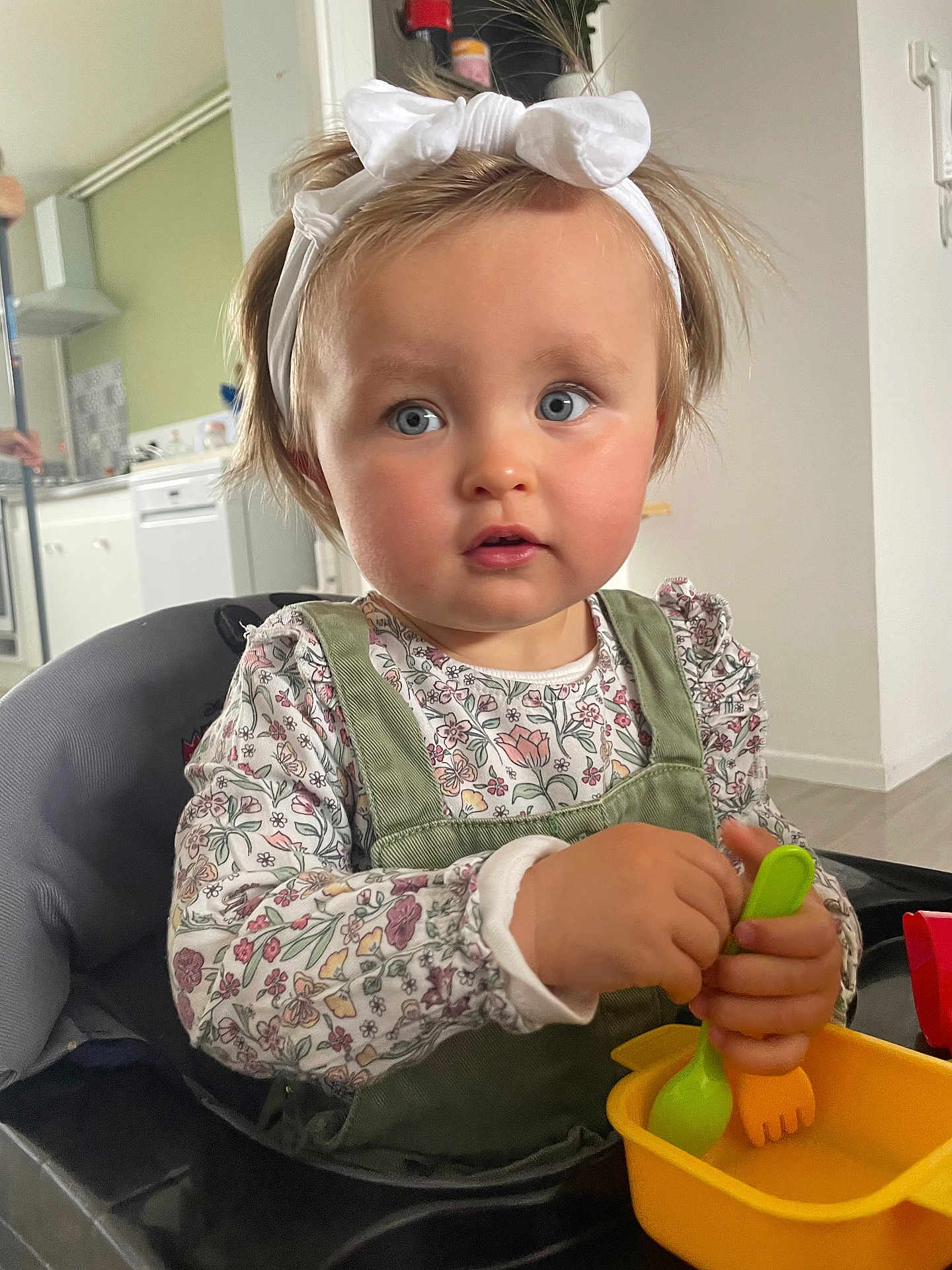Mila a rejoint le concours — aidez-le/la à gagner de superbes lots ! toddler, child, baby, high_chair, kitchen, utensils, yellow_bowl, white_bow, blue_eyes, long_sleeves, green_overalls, curly_hair, indoor, person, cute, feeding, playful, young_child, portrait, table