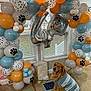 dog, golden_retriever, balloon_arch, birthday, indoor, sweater, paw_print, gift_bag, number_balloon, carpet, window, decorations, party, celebration, blue, orange, white, gray, pet, mat