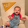armchair, baby, beige, celebration, child, cute, denim_overalls, face, happy, indoor, infant, milestone, pennant, photo, portrait, red_shirt, sitting, smiling, textile, young_child