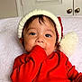 baby, child, red_clothing, hat, hand_in_mouth, blanket, indoors, cute, portrait, infant, holiday, festive, cozy, white_blanket, closeup, smiling_eyes, cute_expression, warm, soft_texture, person