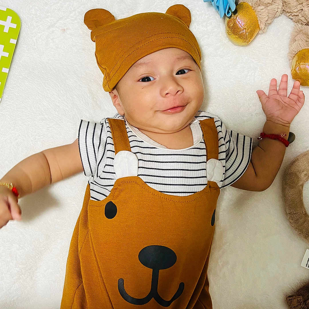 Mathew is registered to the contest to win money with this photo: adorable, baby, bear_outfit, blue_roses, bracelet, casual_clothing, child, cozy, cute, happy, hat, indoors, infant, lying_down, plush_toys, portrait, smiling, soft_surface, striped_shirt, toy_basket