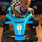 Theo joined the competition — help win amazing prizes! toddler, child, toy_car, blue, smiling, indoor, floor, seat, checkered_pattern, baby_joy, happy, steering_wheel, racing, number_one, person, clothing, striped_shirt, wooden_floor, close_up, playtime