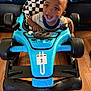 toddler, child, toy_car, blue, smiling, indoor, floor, seat, checkered_pattern, baby_joy, happy, steering_wheel, racing, number_one, person, clothing, striped_shirt, wooden_floor, close_up, playtime