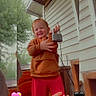 child, toddler, smiling, happy, outdoor, porch, house, siding, hoodie, red_pants, lock, key, holding_object, portrait, standing, heart_stickers, vertical_photo, playful, family, daylight