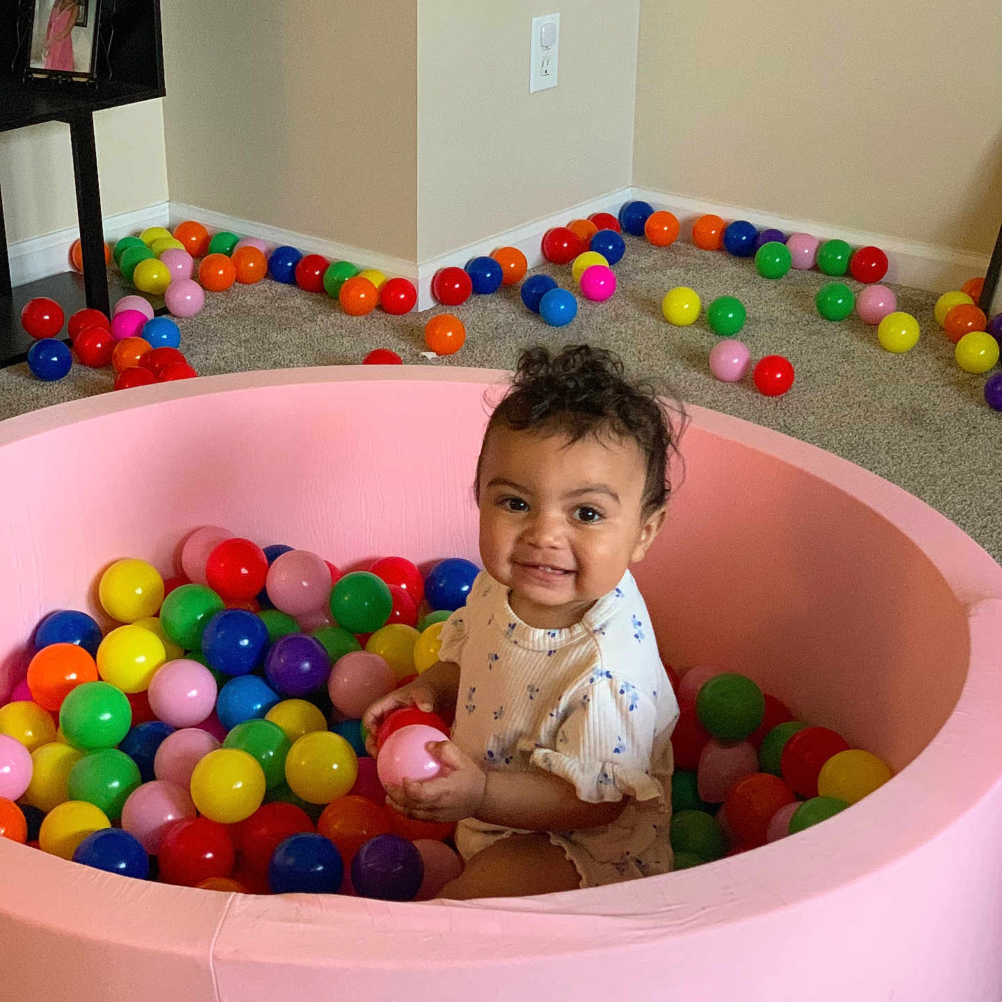 Emberli is registered to the contest to win money with this photo: ball_pit, carpet, child, colorful_balls, curly_hair, furniture, happy, holding, indoor, light_switch, person, photo_frame, pink, plastic_balls, playroom, sitting, smiling, toddler, toy, wall