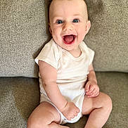 Ezra is registered to the contest to win money with this photo: baby, infant, smiling, happy, onesie, couch, gray, skin, face, blue_eyes, sitting, cute, child, young, indoors, portrait, human, one_person, expression, joy