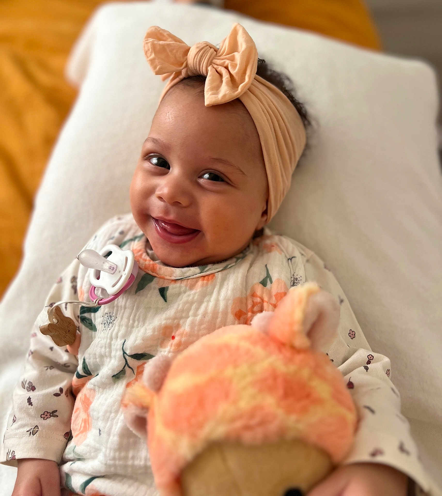 Tahina participe au concours pour gagner de l'argent avec cette photo : baby, smiling, headband, bow, pacifier, toy, plush, floral_clothing, infant, happy, portrait, child, cute, lying_down, blanket, indoor, soft_toy, face, closeup, person