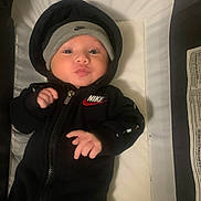 Ace is registered to the contest to win money with this photo: baby, infant, hoodie, nike, black_clothing, beanie, hat, lying_down, soft_surface, indoor, child, cute, puckered_lips, face, hand, young_child, warm_clothing, portrait, person, expression