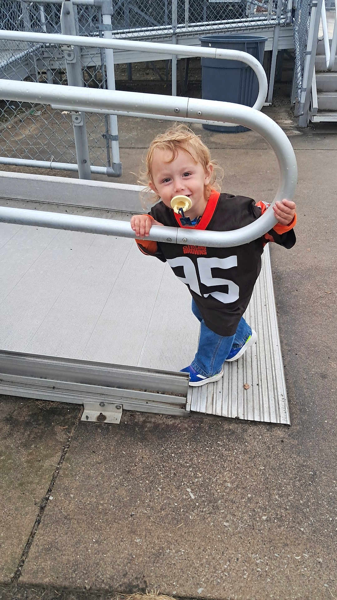 Jackson joined the competition — help win amazing prizes! toddler, child, pacifier, curly_hair, jersey, cleveland_browns, metal_railing, outdoor, concrete, fence, playful, smiling, casual_clothing, blue_jeans, sneakers, young_child, happy, daytime, standing, curly_blonde_hair