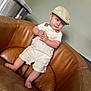 baby, child, infant, overalls, cap, hat, brown_leather_chair, furniture, indoor, curious_expression, hands_clasped, cute, portrait, seated, skin, person, clothing, foot, headwear, home