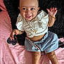 baby, smiling, girl, white_top, blue_skirt, headband, gold_jewelry, bracelet, necklace, earrings, pink_bedspread, heart_pattern, black_pillow, lying_down, happy, cute, infant, person, child, indoors
