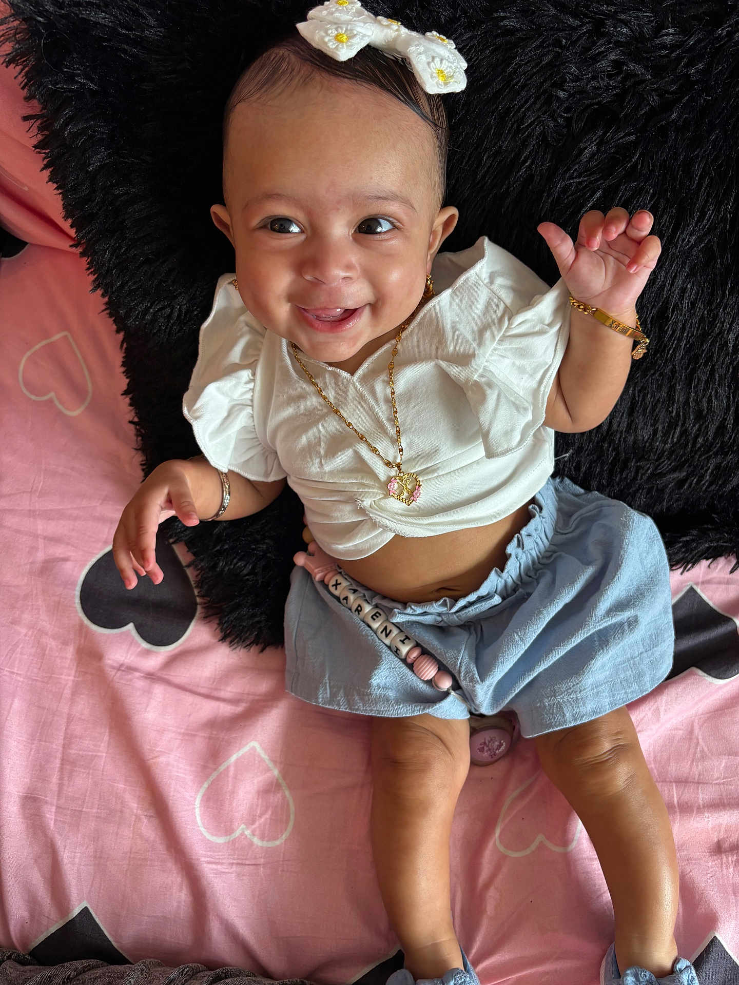 Xareni joined the competition — help win amazing prizes! baby, smiling, girl, white_top, blue_skirt, headband, gold_jewelry, bracelet, necklace, earrings, pink_bedspread, heart_pattern, black_pillow, lying_down, happy, cute, infant, person, child, indoors