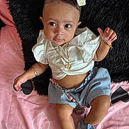 Xareni is registered to the contest to win money with this photo: baby, infant, child, white_bow, white_top, blue_shorts, blue_shoes, gold_jewelry, beaded_chain, pink_blanket, heart_pattern, black_pillow, lying_down, curious_gaze, accessories, cute, indoors, portrait, skin, footwear