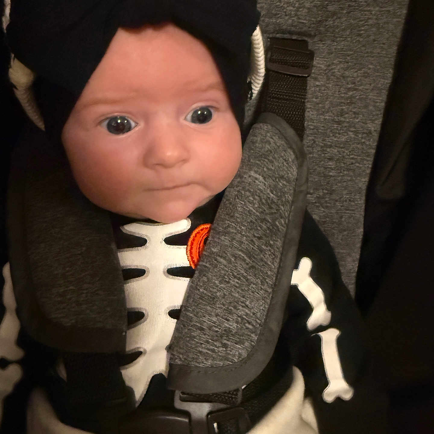 Hannelore is registered to the contest to win money with this photo: baby, baby_clothes, black_bow, blanket, car_seat, child, clothing, costume, cute, expression, face, headwear, indoors, infant, person, portrait, seatbelt, skeleton_costume, straps, wide_eyes