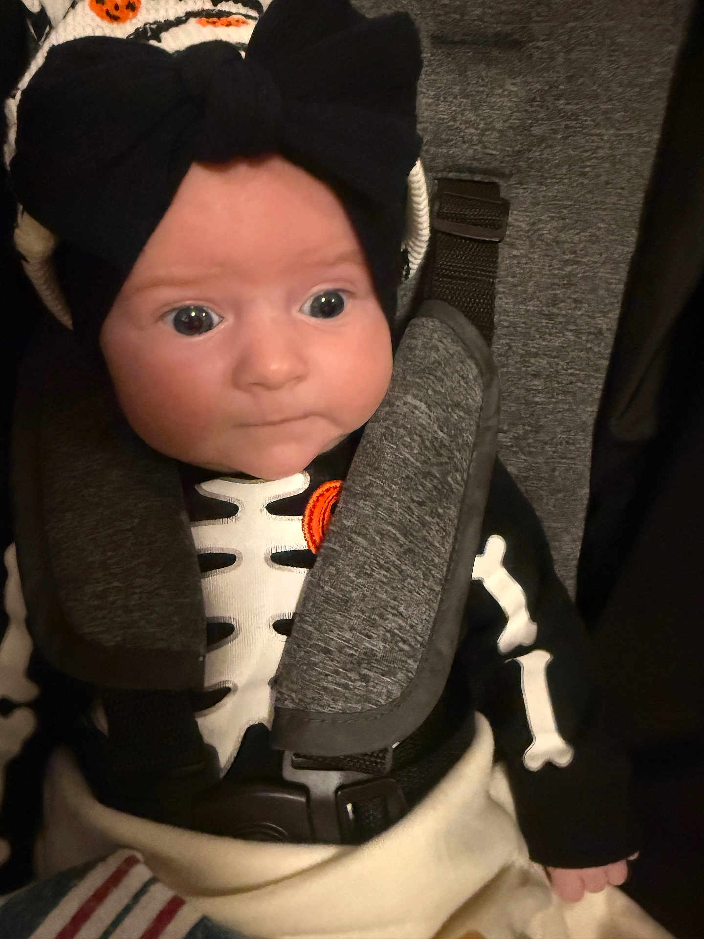Hannelore is registered to the contest to win money with this photo: baby, baby_clothes, black_bow, blanket, car_seat, child, clothing, costume, cute, expression, face, headwear, indoors, infant, person, portrait, seatbelt, skeleton_costume, straps, wide_eyes