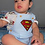 Jose Jr joined the competition — help win amazing prizes! baby, child, superman_logo, clothing, blue_stripes, bed, blanket, sitting, indoor, portrait, cute, infant, person, hand, foot, wall_art, frame, light, expression, young