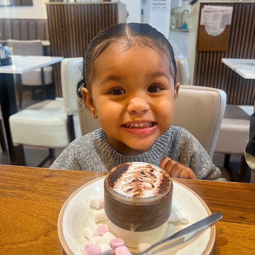 Milaní is registered to the contest to win money with this photo: child, smiling, hot_chocolate, marshmallows, table, chair, cafe, indoor, food, drink, happy, person, dessert, plate, spoon, cozy, young_child, sitting, cute, portrait