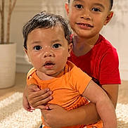 Mason is registered to the contest to win money with this photo: child, baby, sibling, indoor, carpet, red_shirt, orange_outfit, curly_hair, holding, cute, portrait, family, toddler, smiling, happy, home, plant, furniture, candid, warm_light