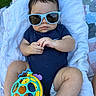 baby, sunglasses, toy, blanket, outdoor, grass, blue_onesie, infant, cute, child, person, legs, hands, face, fashion, relaxing, summer, playful, accessory, cool