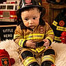 Mason is registered to the contest to win money with this photo: toddler, child, firefighter_costume, helmet, plush_toy, fire_truck_toy, fire_hydrant_toy, sign, indoor, portrait, cute, baby, costume, yellow_stripes, black_boots, fur_rug, brick_wall, firefighter_jacket, sitting, serious_expression