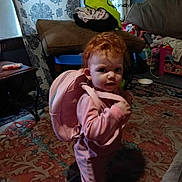 Noel joined the competition — help win amazing prizes! child, toddler, red_hair, pink_backpack, backpack, living_room, rug, couch, curtain, toys, playroom, carpet, pajamas, standing, portrait, indoors, floor, hand, looking_at_camera, curious