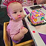 toddler, birthday, cake, frosting, high_chair, headband, messy_face, pink_shirt, celebration, child, party, table, hand, person, food, colorful, indoor, happy, family, seat