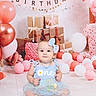 baby, girl, blue_dress, bow, balloons, birthday, gifts, banner, pink, white, floor, celebration, smile, headband, cute, one_year, party, decorations, sitting, indoor