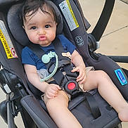 Neil is registered to the contest to win money with this photo: baby, child, infant, stroller, car_seat, pouting, facial_expression, dark_hair, blue_shirt, shorts, pacifier, teether, harness, safety_buckle, portrait, sitting, indoor, tile_floor, photograph, cute